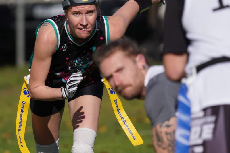 A flag football player in a black and teal kit, with gloved hands and a yellow flag belt, faces the camera