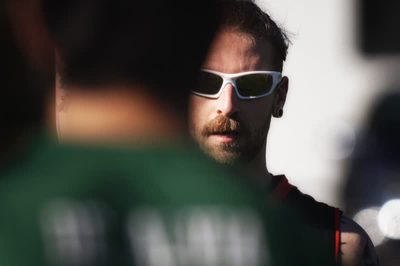 A bearded player in white-framed sunglasses looks out from behind a blurred teammate in a green shirt
