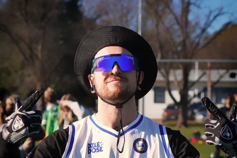 A flag football player in a bucket hat and blue wraparound sunglasses raises gloved hands to the camera