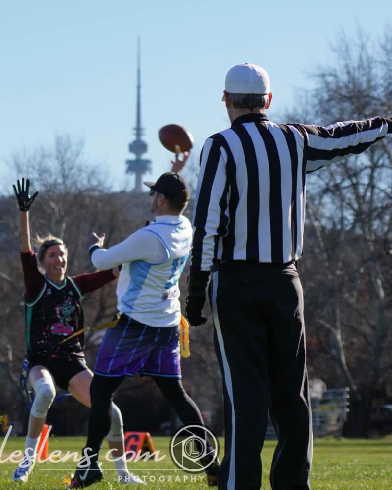 A flag football quarterback throws past a referee, Telstra Tower behind, as posted by American Football ACT