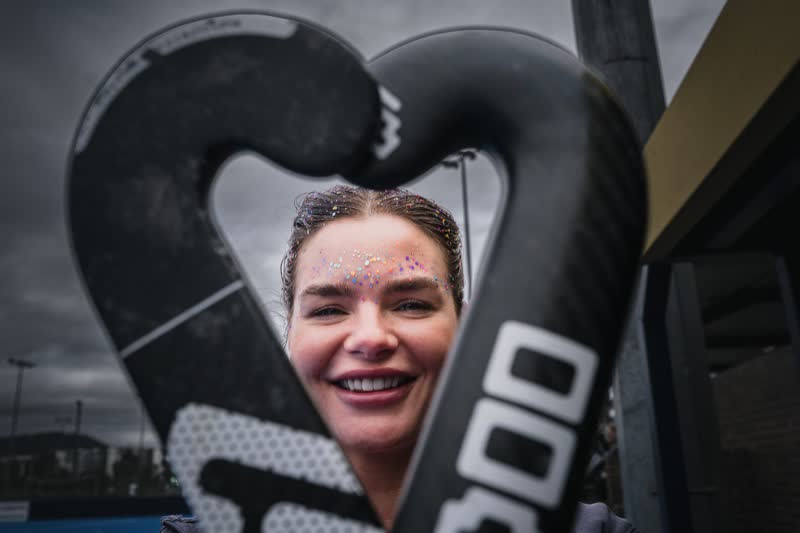 Hockey ACT Pride Round: a smiling player with glitter on her face frames herself between two hockey sticks held in a heart shape