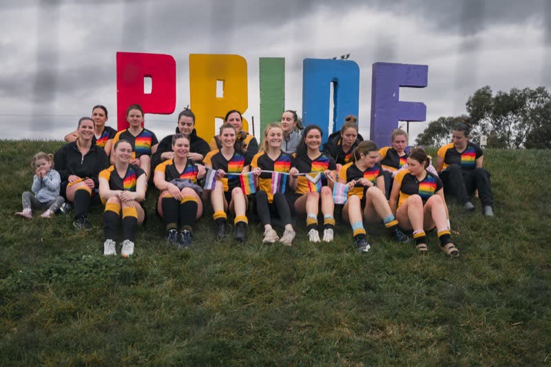 Hockey ACT Pride Round: a team in rainbow-trimmed shirts sits together on the grass in front of a large PRIDE sign