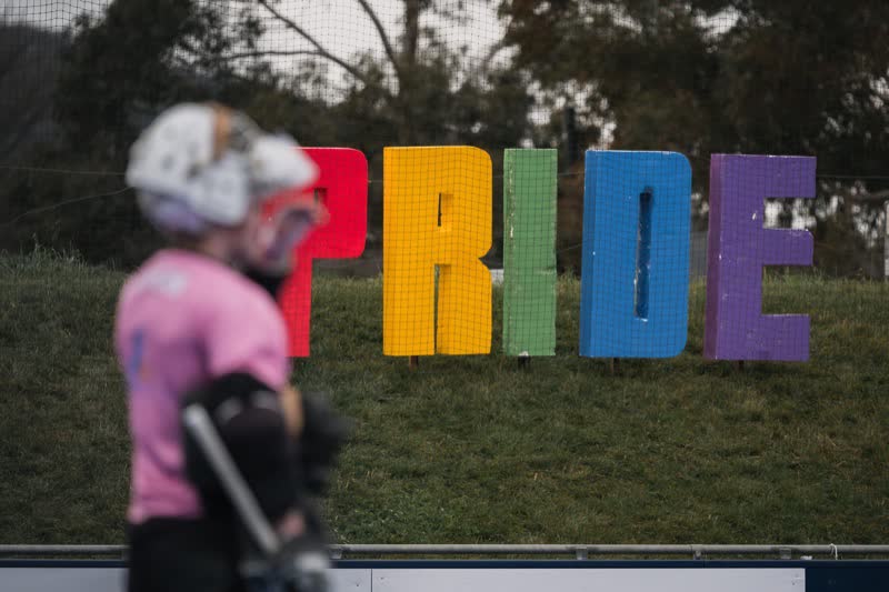 Hockey ACT Pride Round: a blurred player in a pink shirt and helmet in the foreground, the rainbow PRIDE letters on the bank behind